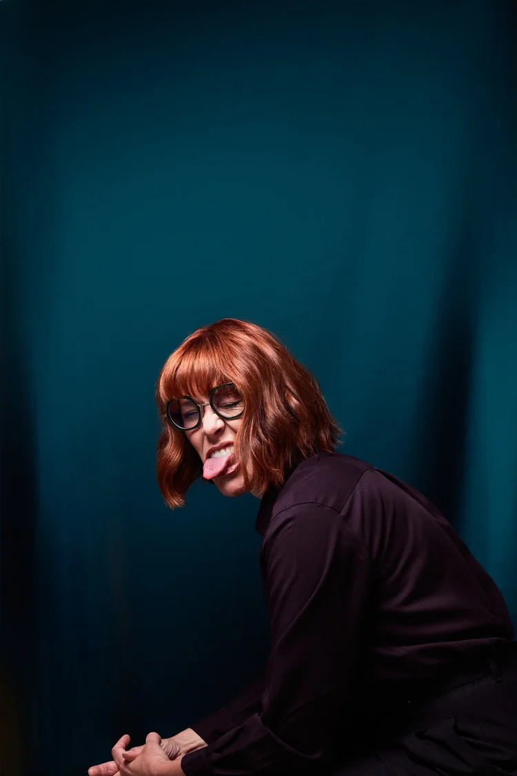 Une femme avec des cheveux roux, portant des lunettes rondes noires, fait semblant de tirer la langue et de grimacer. Elle porte une chemise noire et est assise contre un fond bleu foncé.