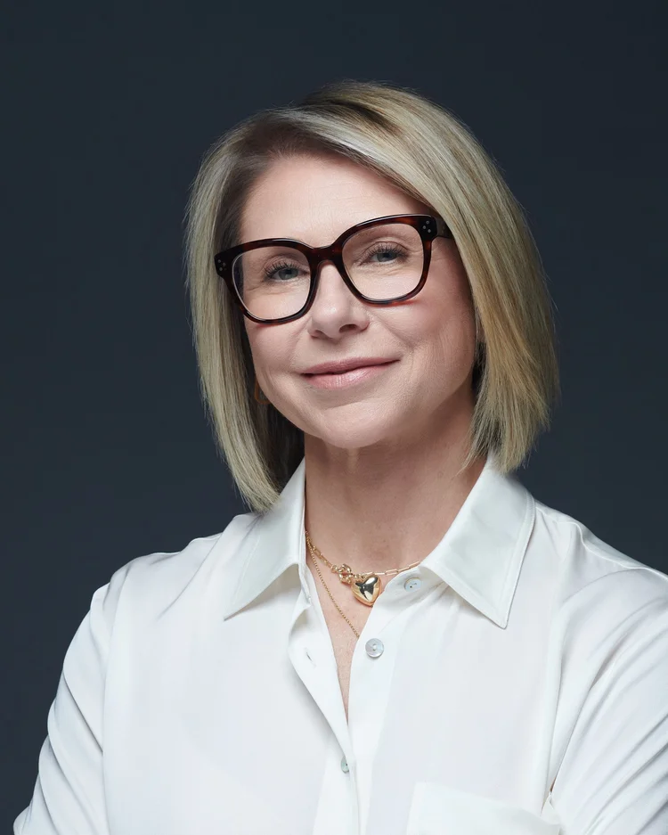 Femme aux cheveux blonds mi-longs portant de grandes lunettes noires et une chemise blanche, souriante, sur fond gris foncé.