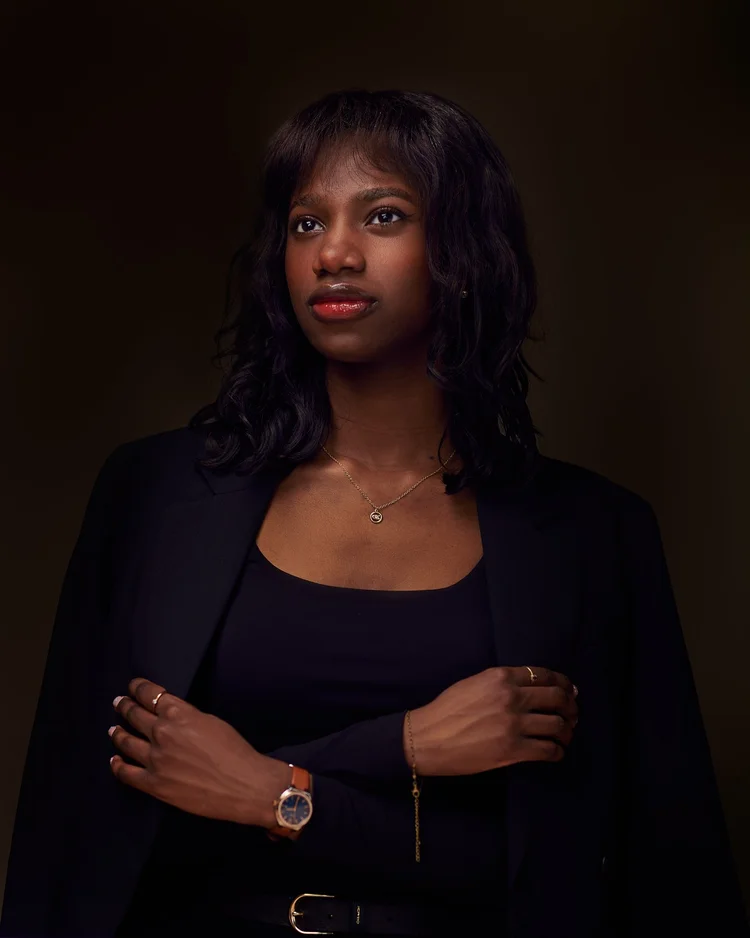 Portrait d'une femme aux cheveux bouclés portant un tailleur noir, une montre dorée et un collier fin, bras croisés, sur fond brun sombre.