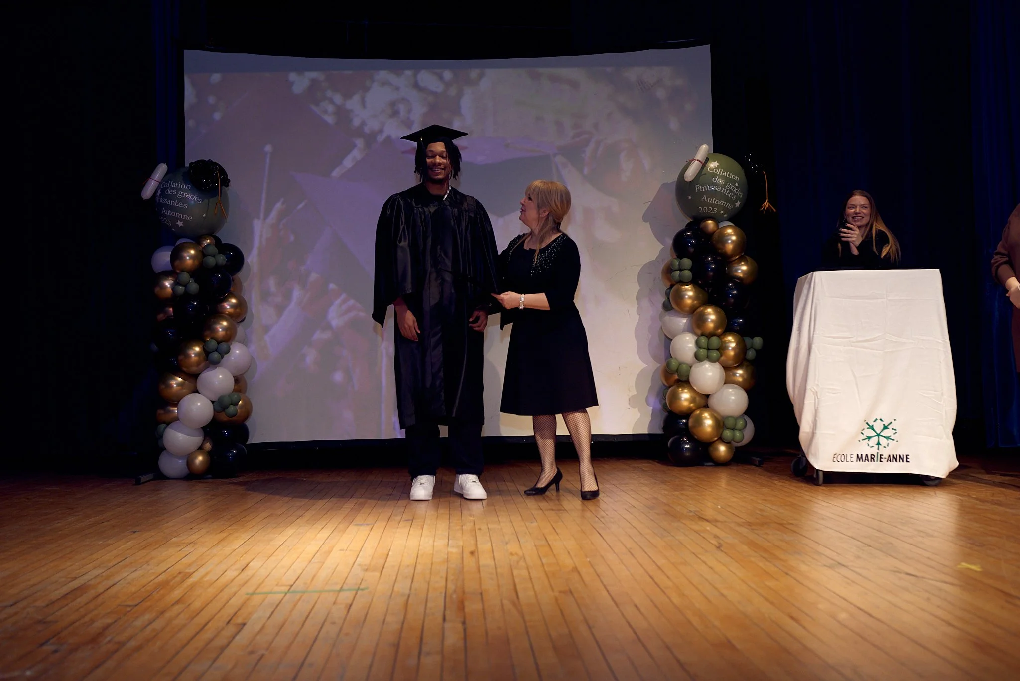 Cérémonie de remise de diplôme dans une salle de spectacle avec un décor de ballons, une personne en costume de fête recevant un diplôme, et une autre personne en robe. Présence d'un écran en arrière-plan et de personnes à côté, une femme à droite qu