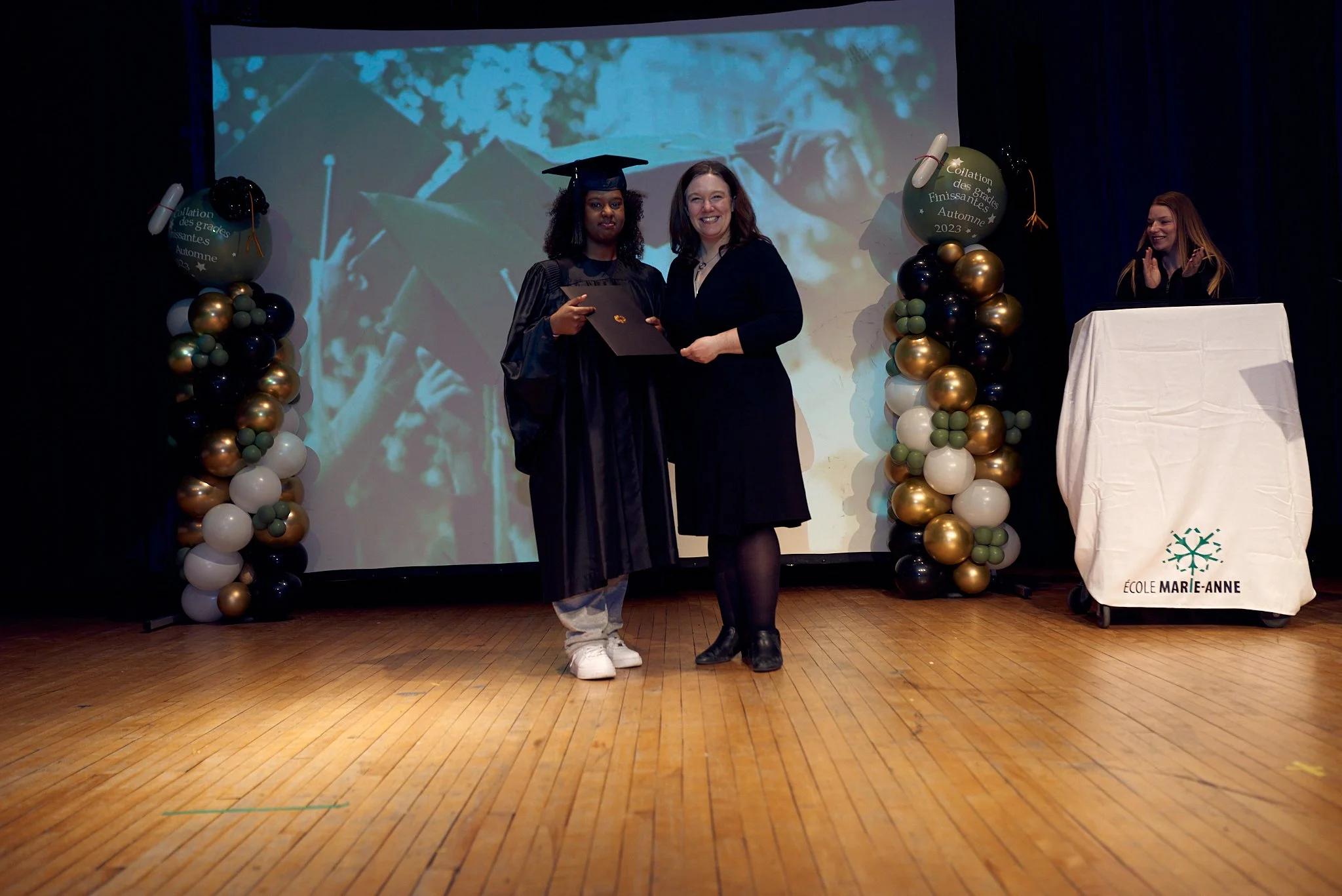 Une cérémonie de remise de diplôme à l'école Marie-Anne, avec une femme diplômée en costume de graduation recevant un diplôme d'une autre femme. Il y a des ballons décoratifs et une personne à un pupitre sur le côté.