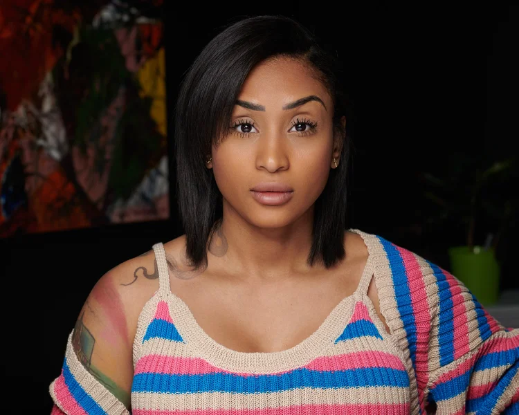 Un portrait d'une femme aux cheveux noirs, portant un pull rayé de couleurs rose, bleu et beige, avec un tatouage sur l'épaule gauche. Fond sombre, avec une peinture abstraite floue à gauche.