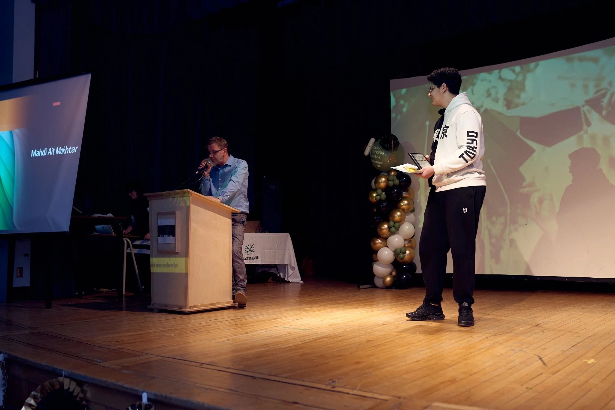 Une scène de présentation ou de discours dans une salle de théâtre ou d'auditorium. Deux hommes sont visibles : l'un derrière un pupitre et l'autre debout avec un micro et un tablette. Il y a un grand écran derrière eux, avec des ballons décoratifs à