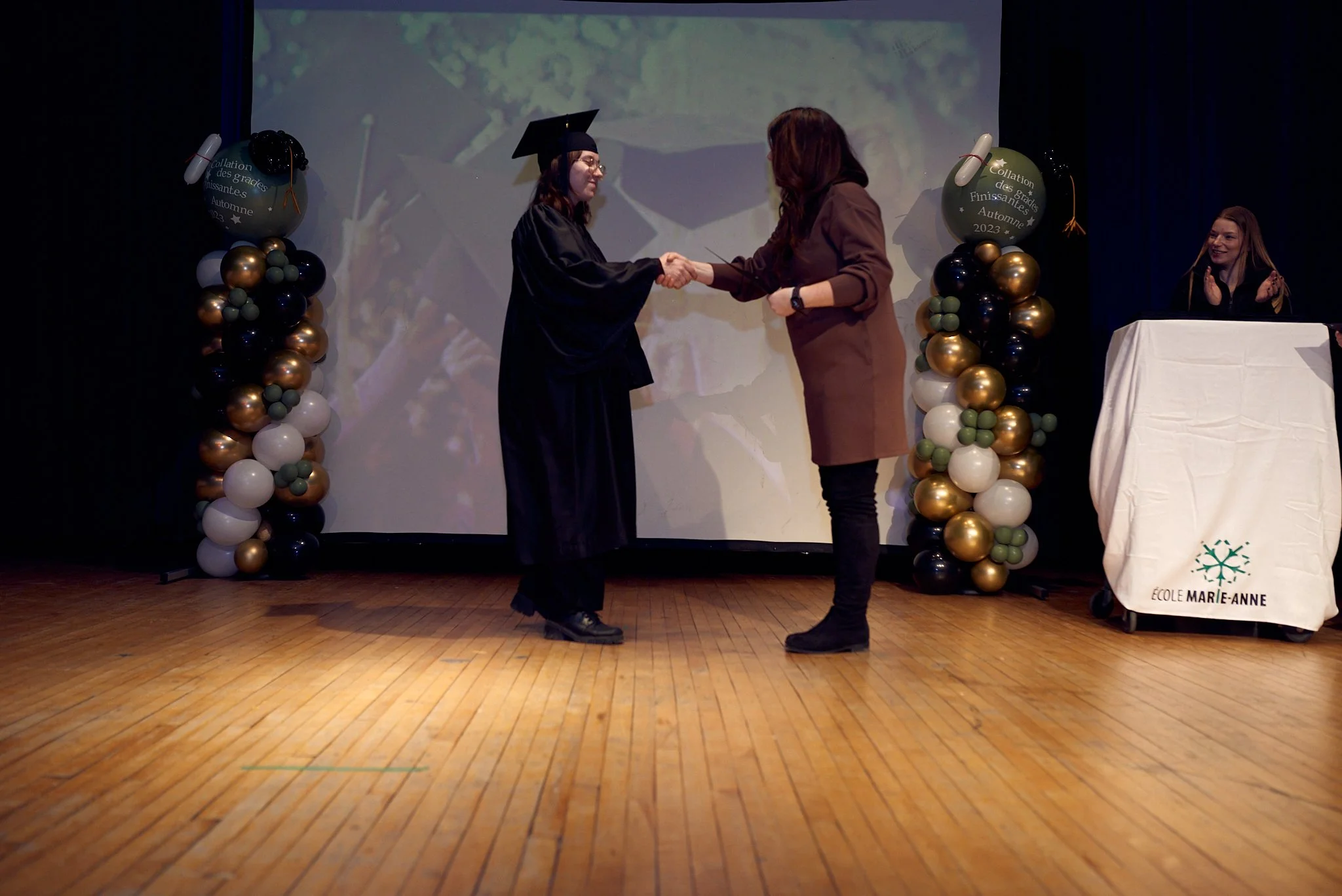 Une femme en robe de graduation serre la main d'une autre femme lors de la cérémonie de remise des diplômes sur scène, avec des décorations de ballons dorés, noirs et blancs.