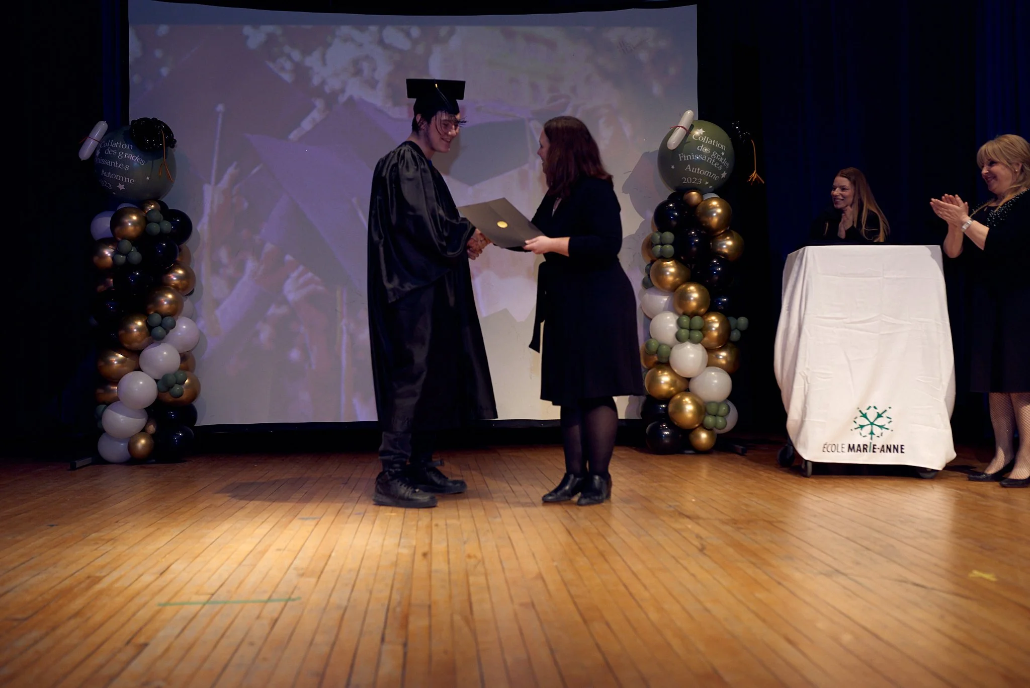 Une cérémonie de remise de diplôme à une graduation avec deux femmes échangeant un diplôme sur scène, entourées de balloons et un écran en arrière-plan. Une femme en robes de graduation en noir et un chapeau mortarboard recevant le diplôme, lors d'un