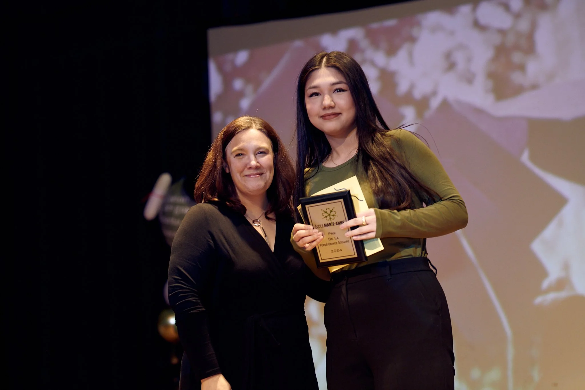 Une jeune femme reçoit un prix lors d'une cérémonie sur scène, tenue en main, en compagnie d'une femme plus âgée, toutes deux souriantes.