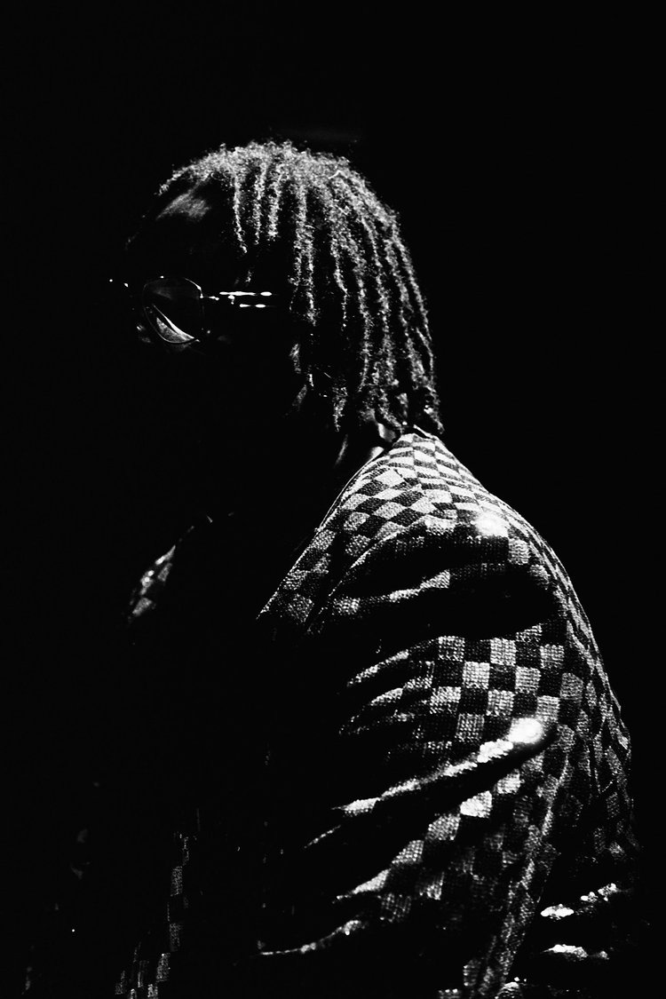 Un portrait en noir et blanc d'une personne portant des lunettes, avec des cheveux torsadés, vêtue d'une chemise à carreaux, face partiellement tournée et dans un éclairage sombre.