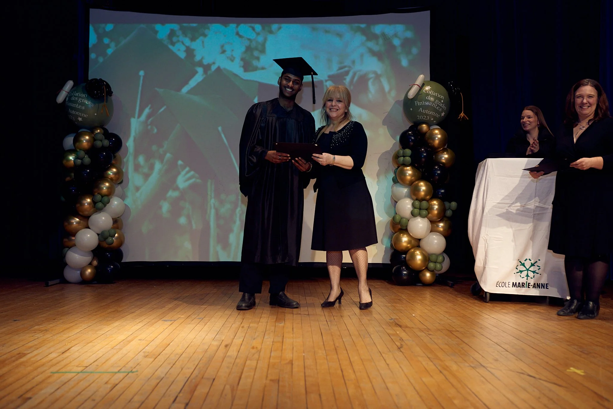 Un diploma de fin d'études est remis lors d'une cérémonie de remise de diplômes à l'école Marie-Anne. Un jeune diplômé en robe de graduation et toque, souriant, reçoit le diplôme d'une femme en robe noire. Deux autres femmes se tiennent à droite près