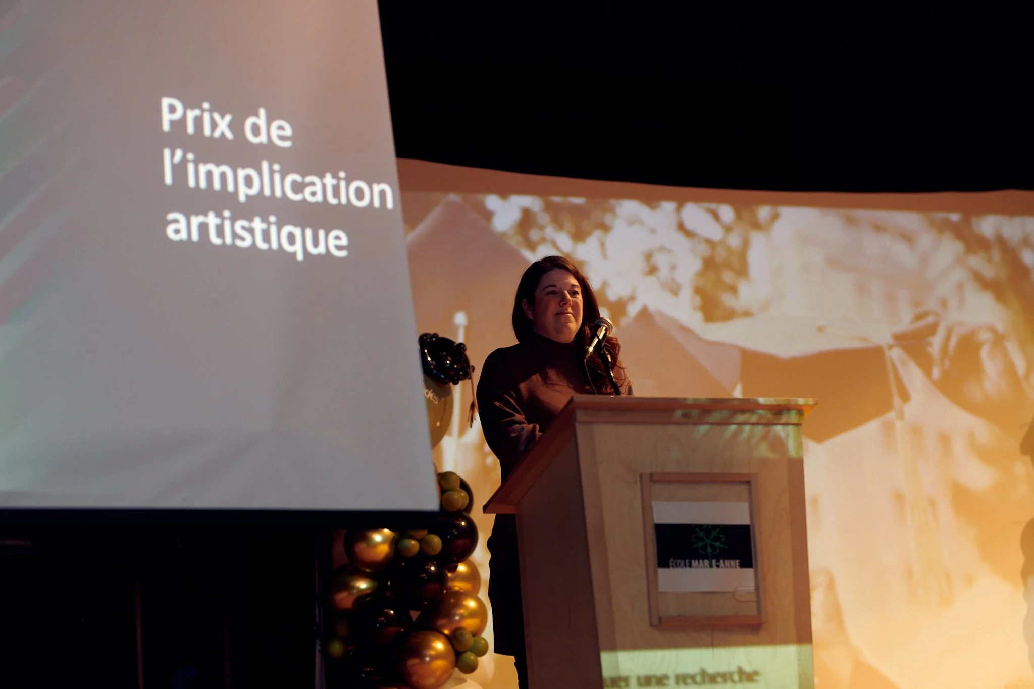 Une femme donne une présentation à un podium avec une grande projection en arrière-plan et une décoration de ballons dorés et noirs.