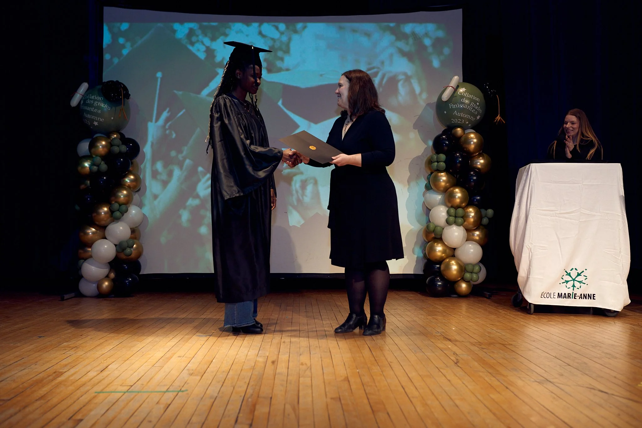 Une cérémonie de remise de diplôme à une école appelée École Marie-Anne, avec deux femmes échangeant un certificat sur scène. Une femme en robe de graduation en noir et une autre en tenue professionnelle. Décorations de ballons et une personne souria