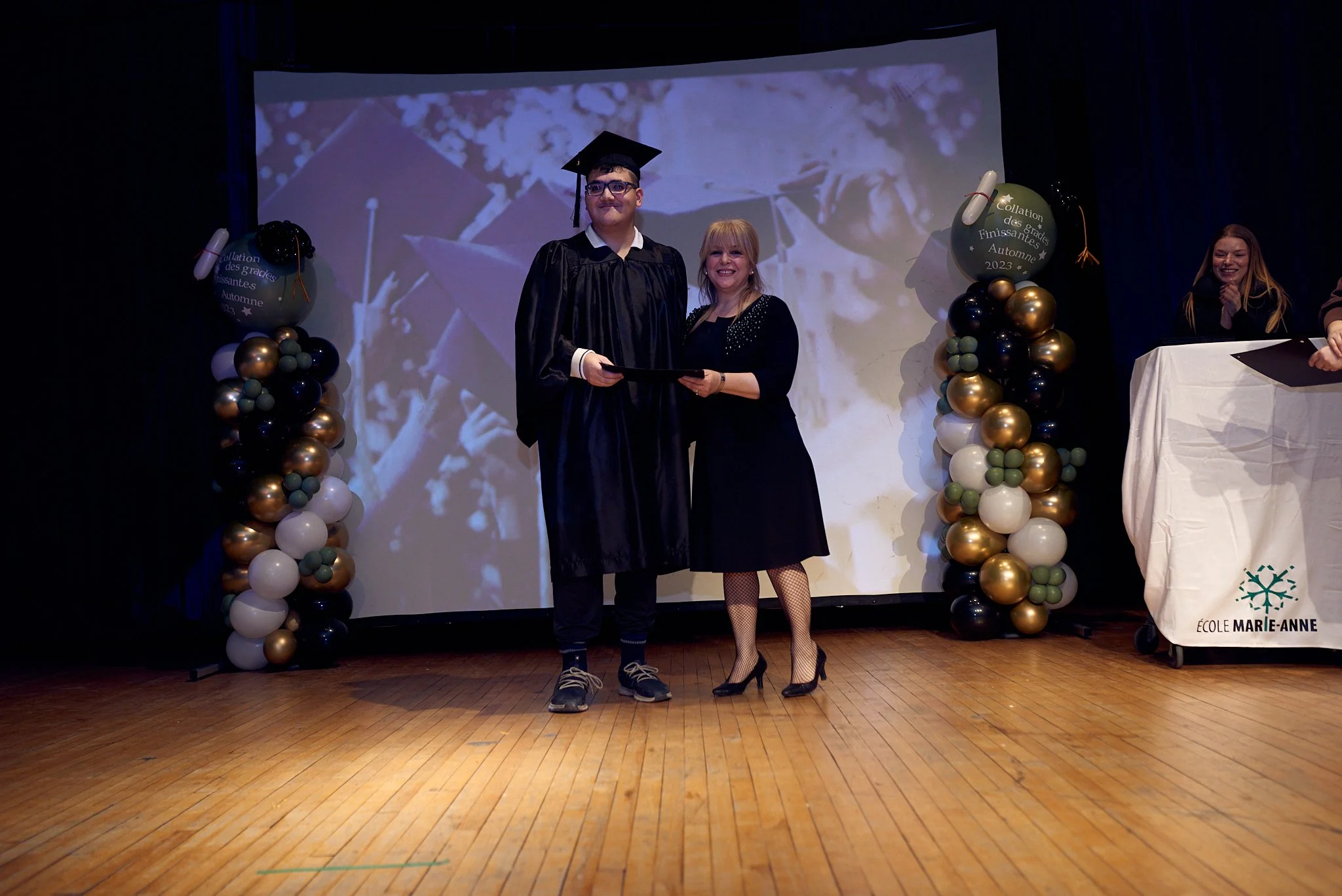 Un jeune homme en robe de graduation et une femme souriante lors d'une remise de diplôme dans une cérémonie. Ils se tiennent sur scène, entourés de décorations de ballons et d'un grand écran en arrière-plan affichant une image floue.