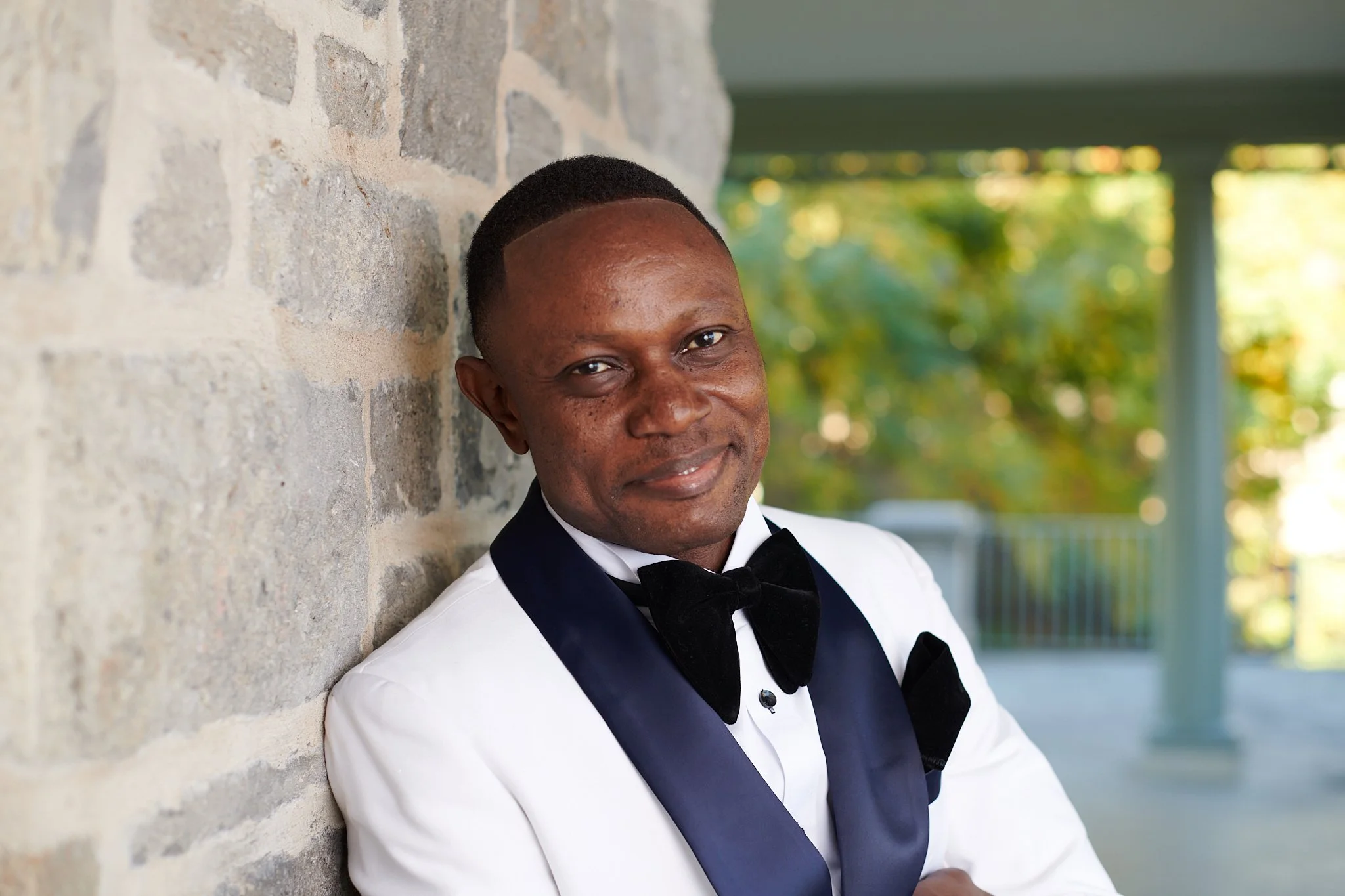 Homme en smoking blanc avec nœud papillon noir, assis contre un mur en pierre, souriant à la caméra, fond flou avec des arbres verts.