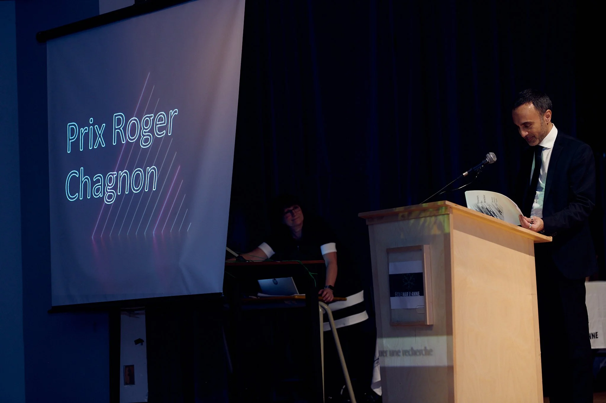 Un homme en costume parle lors d'une cérémonie avec un panneau indiquant "Prix Roger Chagnon".