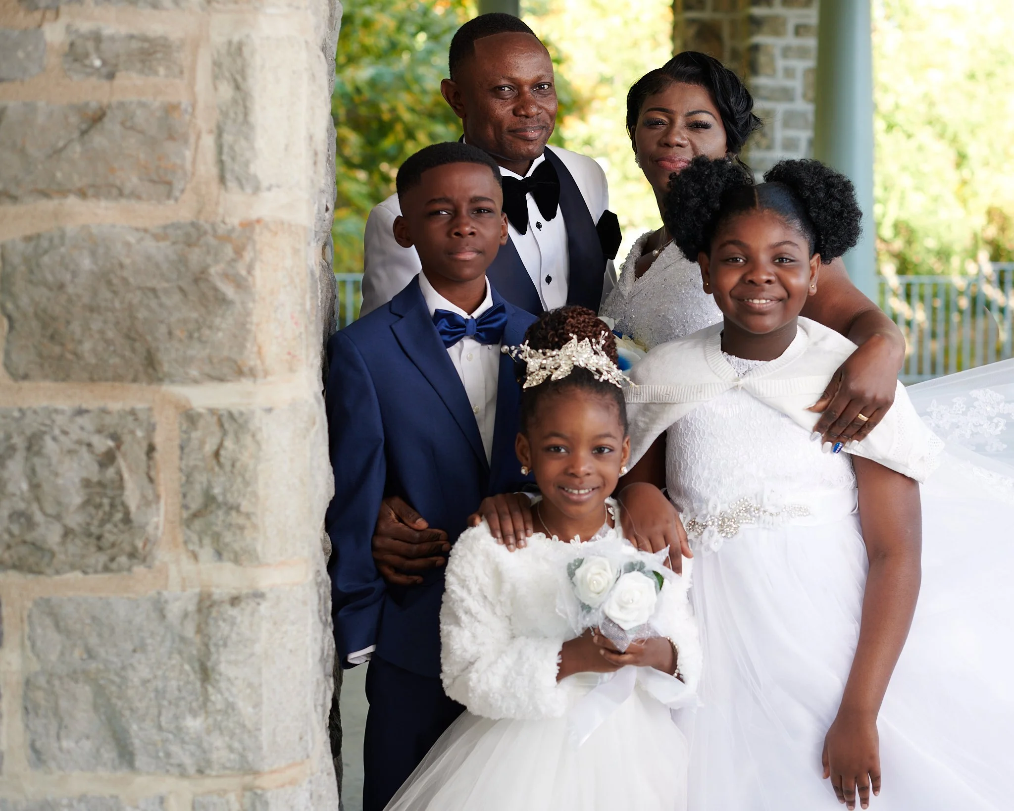 Groupe de six personnes lors d'un mariage, dont deux adultes et quatre enfants, posant devant une structure en pierre avec un environnement extérieur vert en arrière-plan, tous élégamment habillés, avec la petite fille tenant un bouquet de fleurs.
