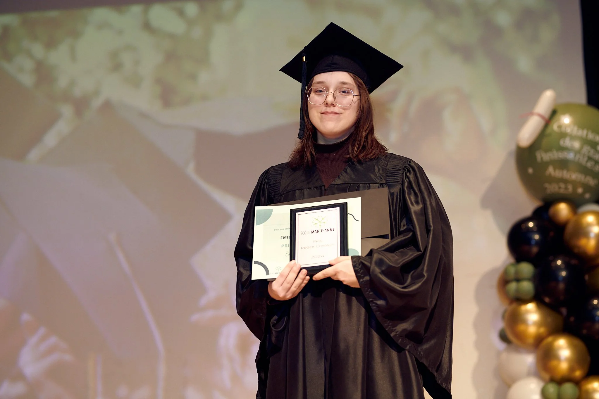 Jeune femme en robe de graduation noire, portant un chapeau carré, tenant un certificat d'obtention de diplôme sur scène, avec un mur de décor en arrière-plan et un ballon de fête coloré à droite.