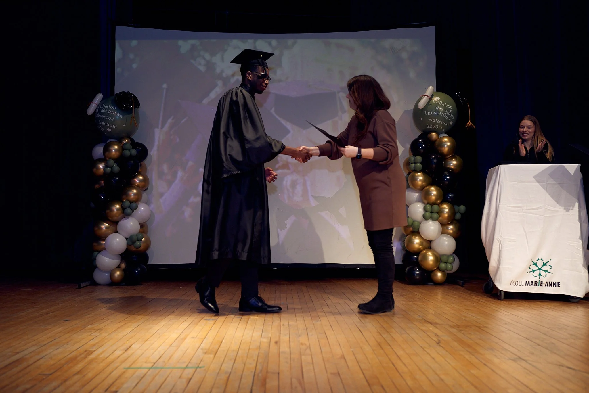 Jeune homme en graduation en robe et toque recevant son diplôme lors d'une cérémonie, échanger une poignée de main avec une femme sur scène, décorée avec des ballons et une bannière "École Marie-Anne".