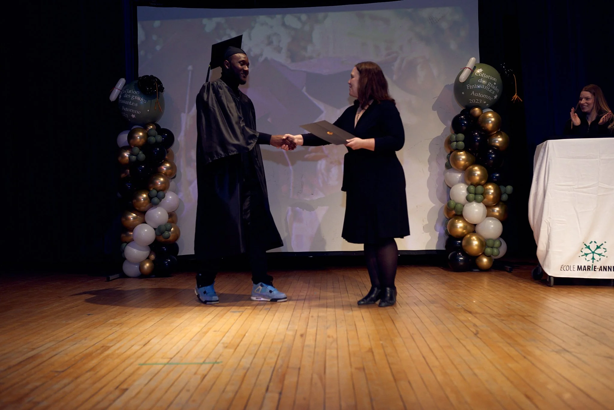 Un étudiant en tenue de graduation recevant un diplôme lors d'une cérémonie sur scène, décorée avec des ballons noirs, or, blanc et vert, sous un fond projection vidéo.