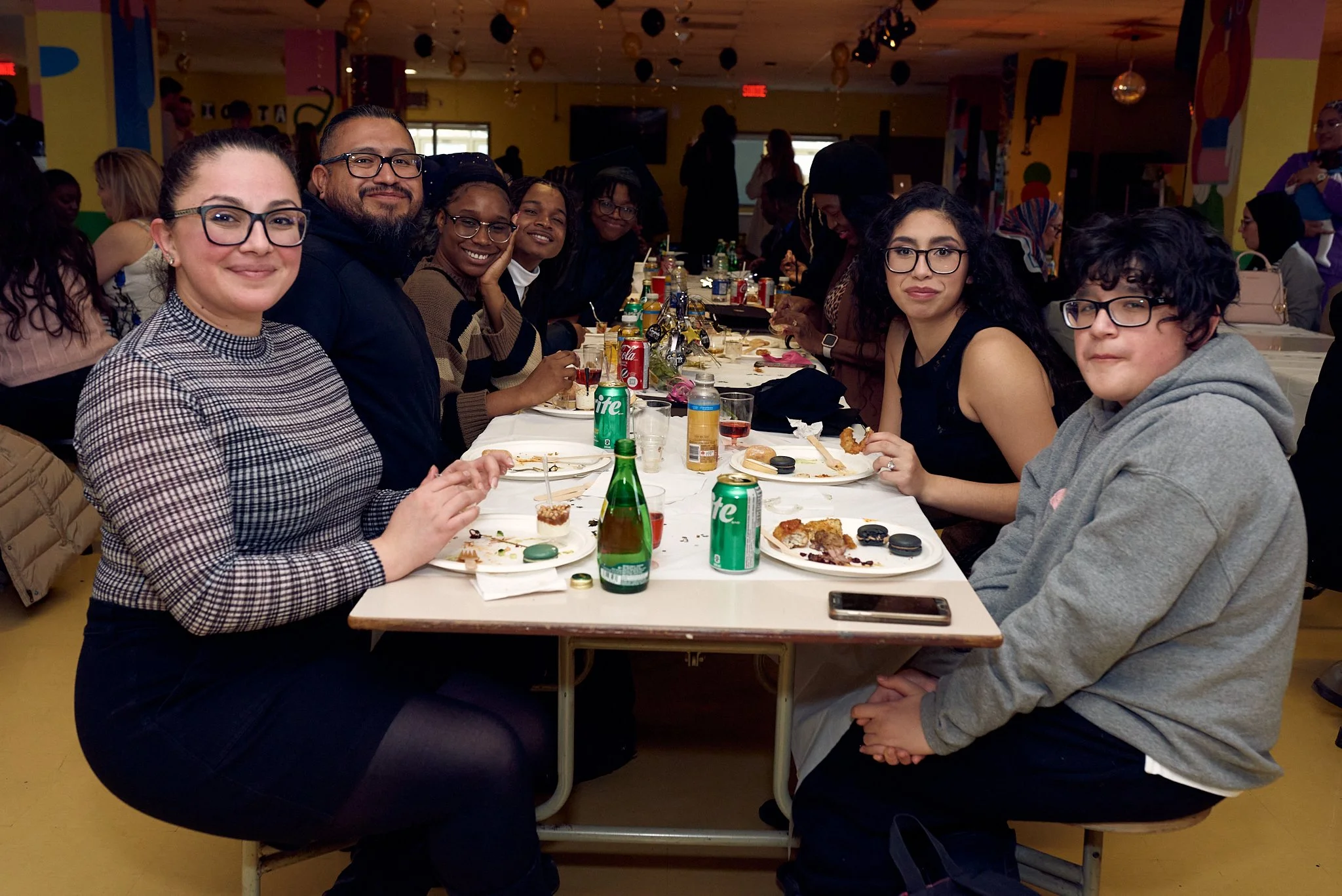 Groupe de personnes assises autour d'une table lors d'une fête ou d'un repas convivial, avec boissons et nourriture sur la table.