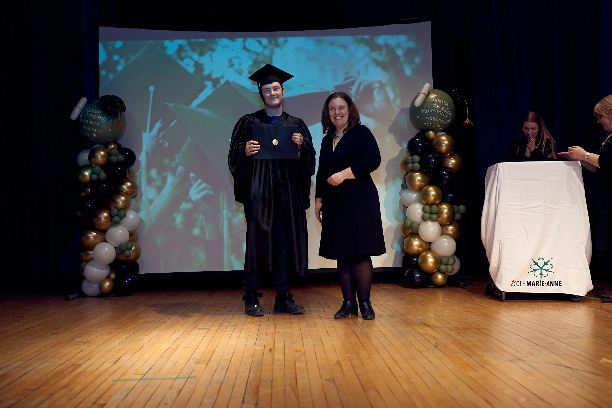 Étudiant en costume de graduation recevant son diplôme lors de la remise de diplômes à l'école Marie-Anne, avec une personne officielle à ses côtés, dans une salle décorée avec des ballons.