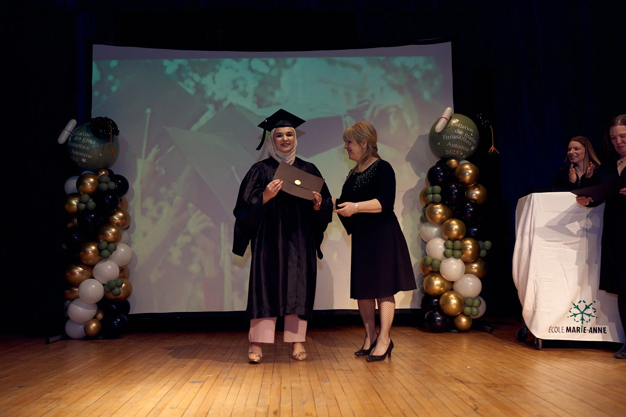 Une cérémonie de remise de diplôme dans une école, avec une étudiante en robe de graduation recevant son diplôme de la part d'une femme, sur scène décorée avec des ballons de couleur or, blanc et vert, et un grand écran en arrière-plan affichant une 