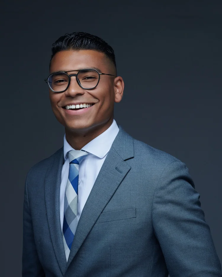 Homme souriant portant des lunettes, complet gris et cravate à motifs, portrait corporatif sur fond gris uni.