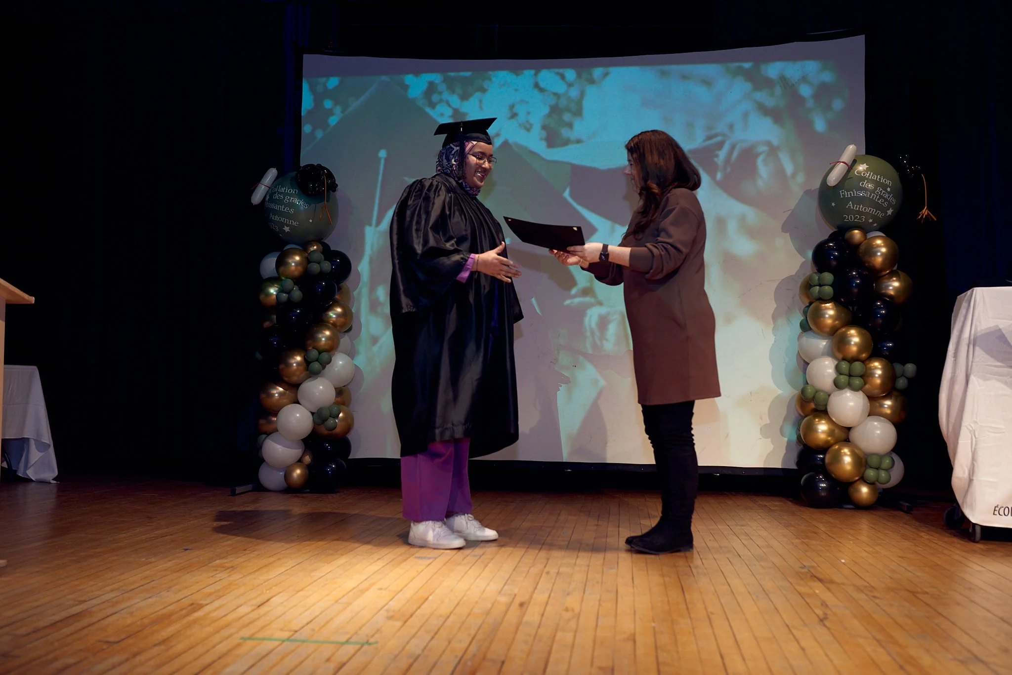 Une personne en robe de graduation reçoit un diplôme lors d'une cérémonie sur scène, avec des décorations de ballons
