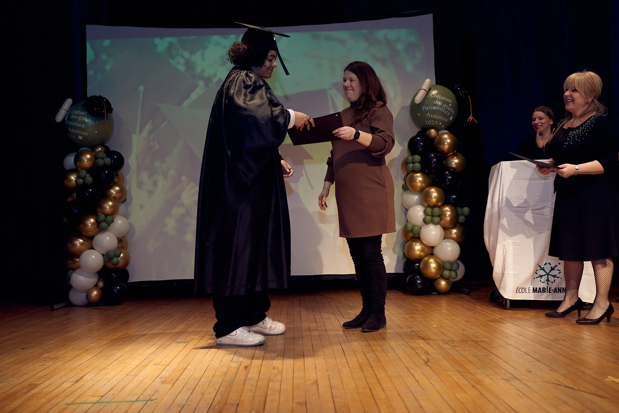 Une cérémonie de remise de diplôme scolaire avec une étudiante en robes de graduation, recevant son diplôme d'une femme lors d'une cérémonie académique, décorée de ballons noirs, or et blancs.