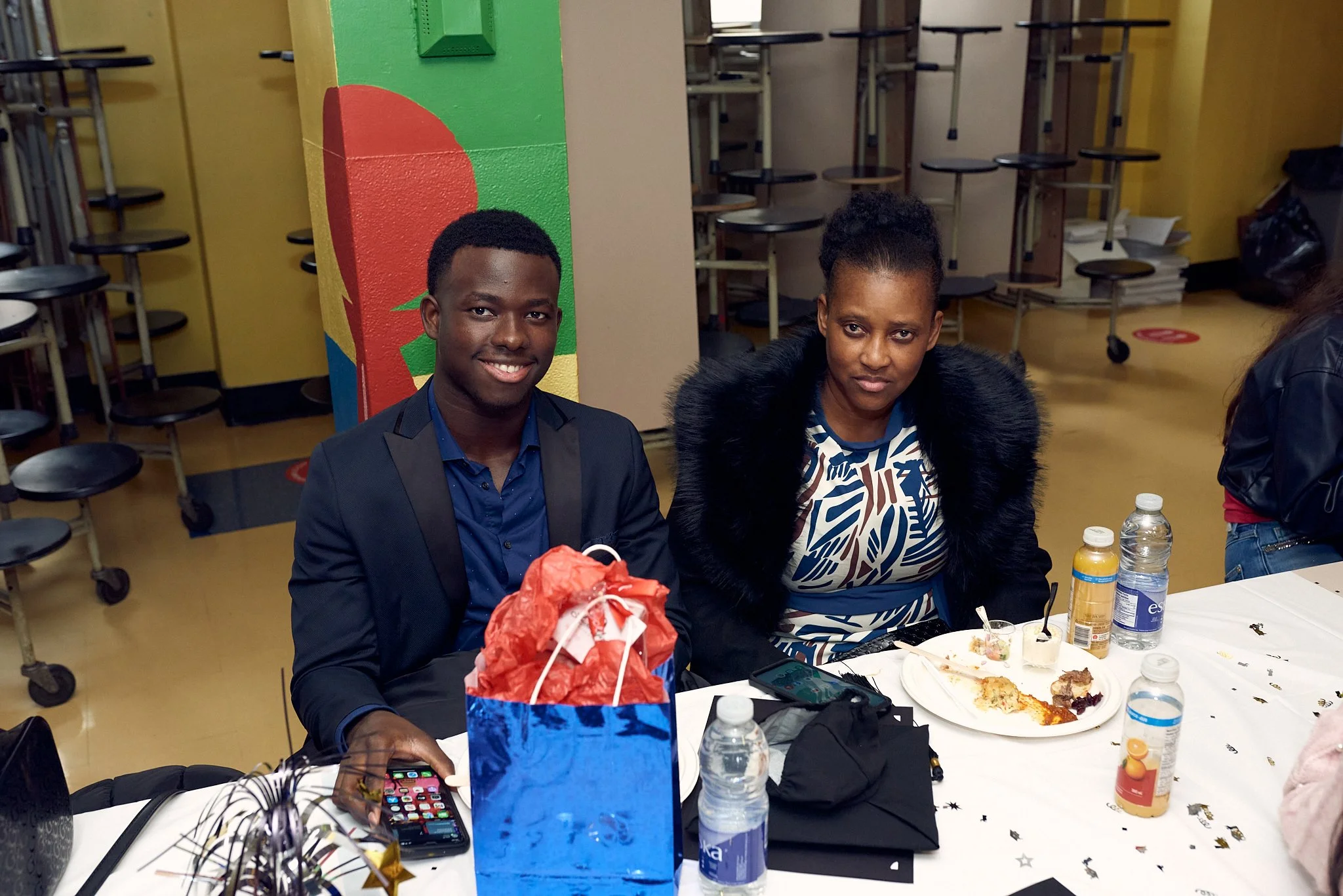 Deux personnes assises à une table lors d'une fête, avec des bouteilles d'eau, des plats de nourriture et des cadeaux.