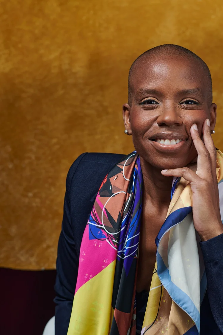 Femme au crâne rasé souriant largement, la main sur la joue, portant un foulard en soie coloré et un blazer marine, fond doré texturé.