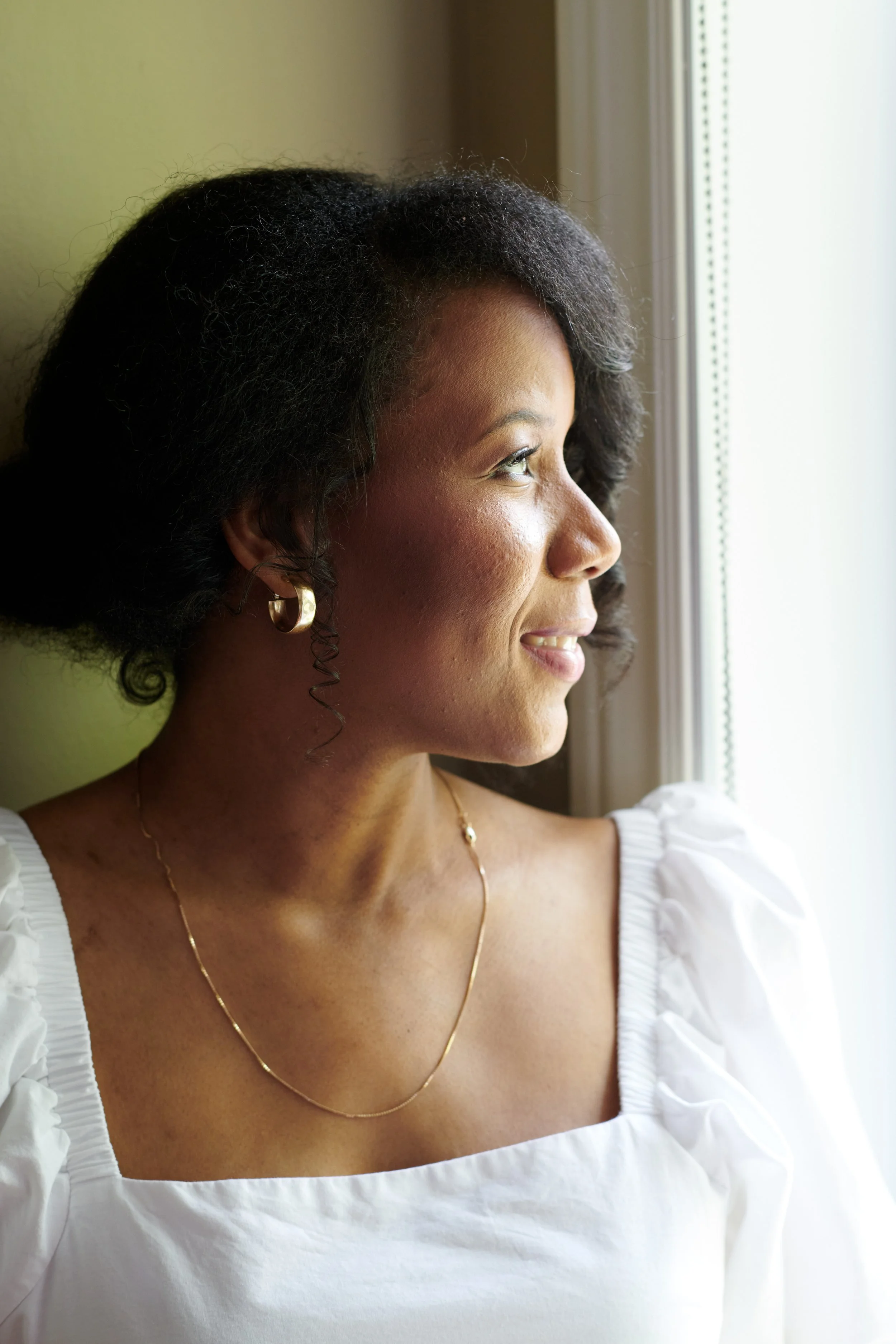 Femme noire regardant par une fenêtre, portant des boucles d'oreilles en or et un collier, avec une blouse blanche