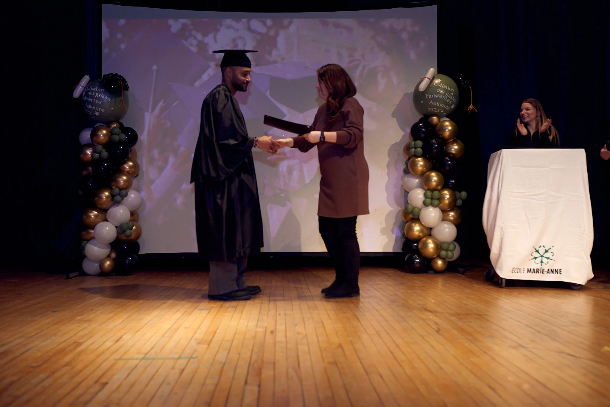 Une cérémonie de remise de diplôme sur scène avec un diplômé en robe et bonnets de graduation recevant un diplôme d'une femme lors d'un événement scolaire, décoré de ballons et d'un fond d'écran de graduation.