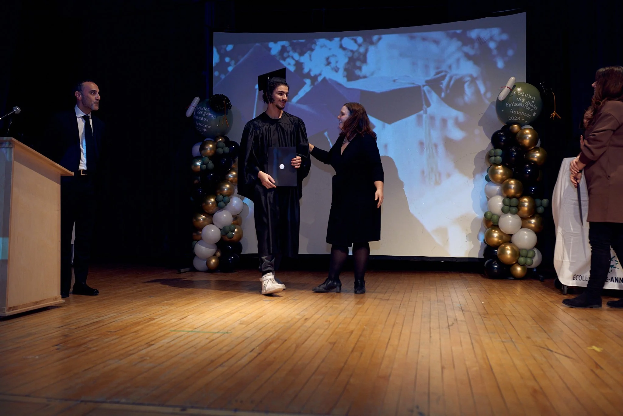 Cérémonie de remise de diplôme lors d'une cérémonie de fin d'études, avec un diplômé en robe de graduation en train de recevoir son diplôme sur scène, décorée de ballons et d'une projection en arrière-plan.