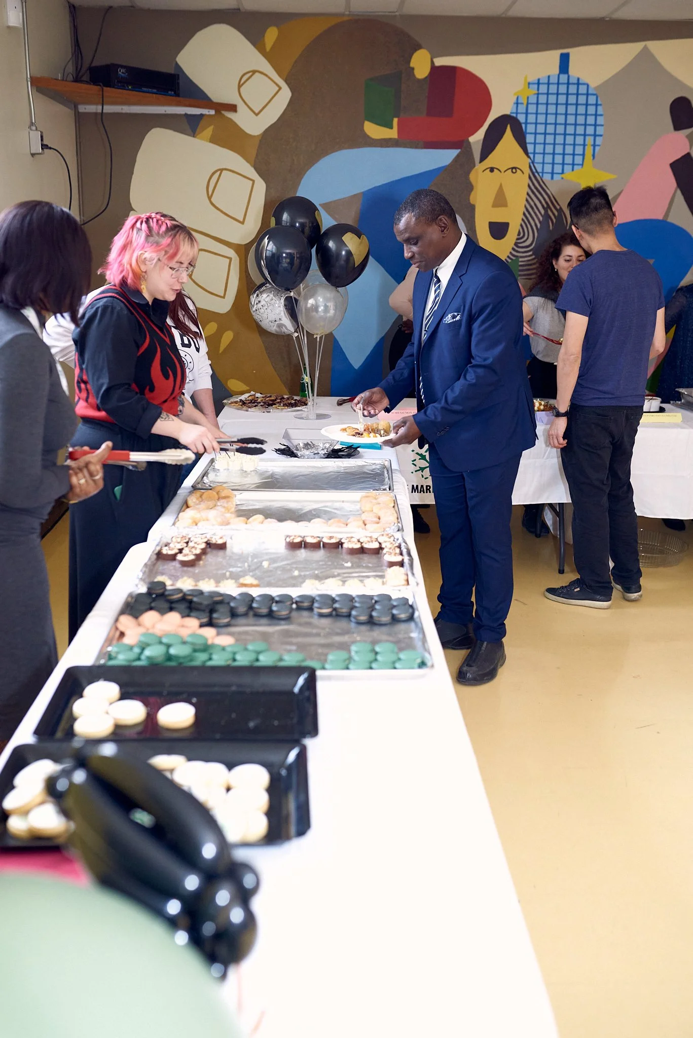 Une table avec des cupcakes colorés lors d'une célébration, des personnes en train de se servir, et un mur peint avec des formes géométriques et des visages stylisés en arrière-plan.