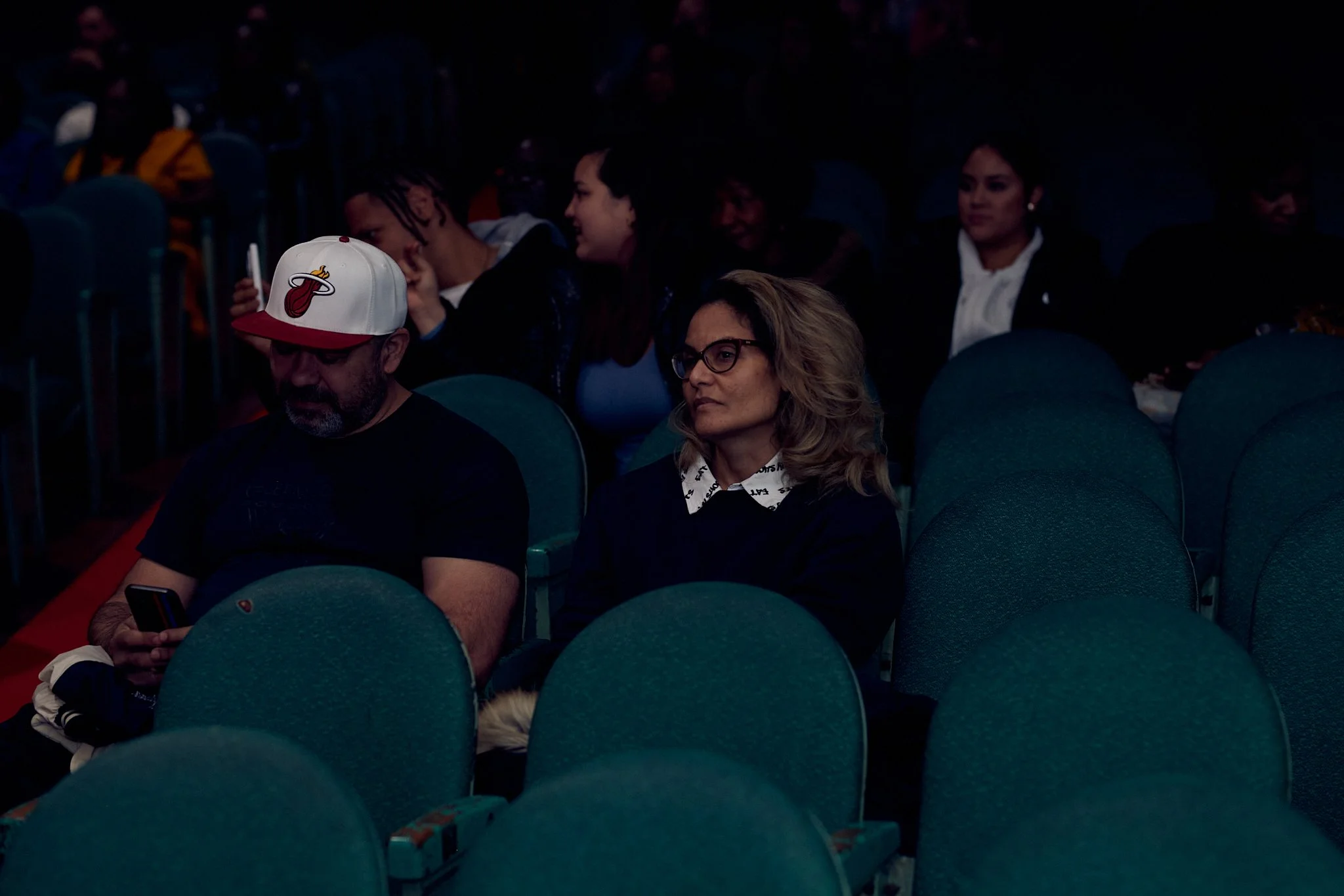 Des personnes assises dans une salle sombre, dont un homme portant un chapeau de baseball Miami Heat et utilisant son téléphone, et une femme avec des lunettes et des cheveux bouclés assise à côté de lui.