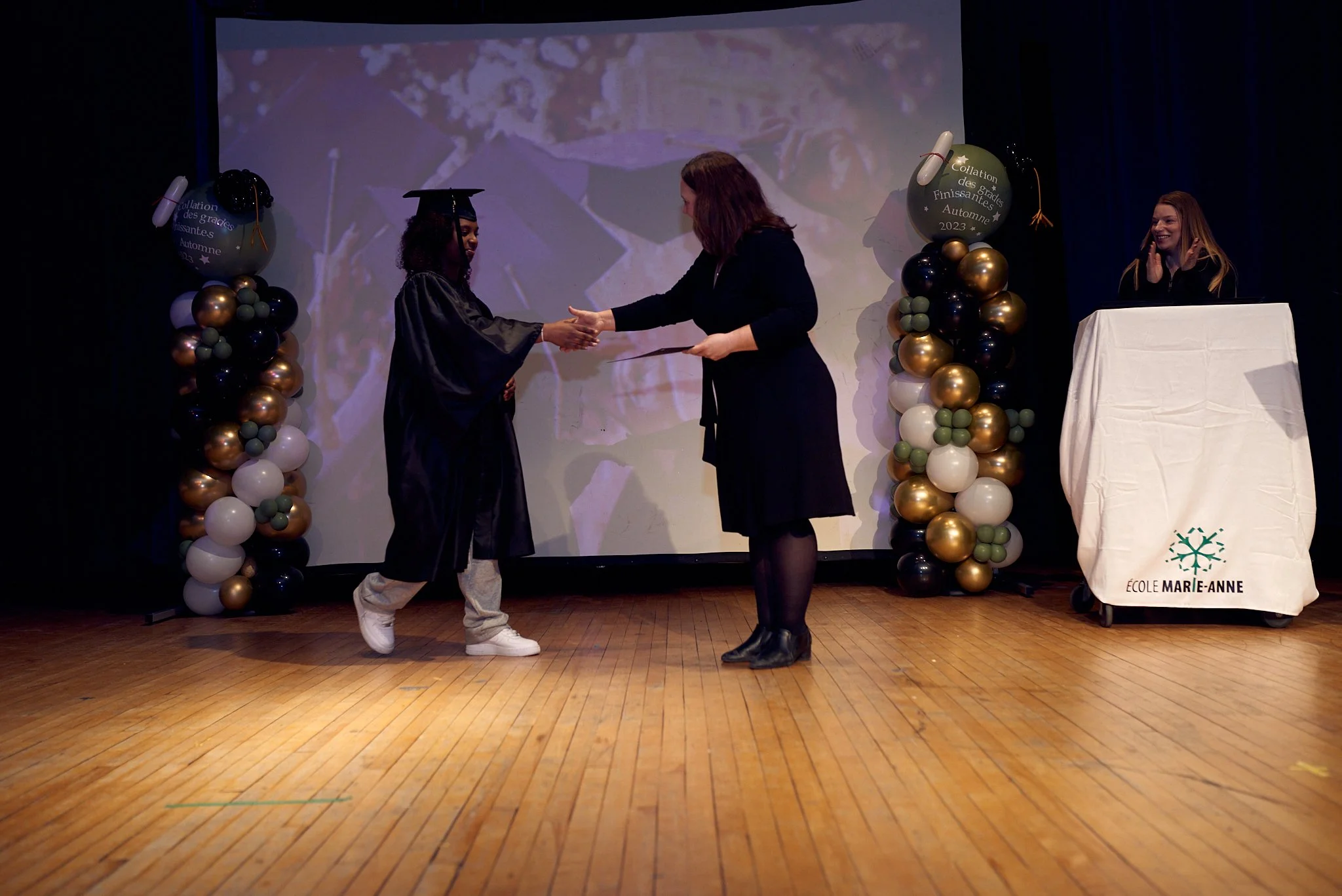 Une cérémonie de remise de diplôme sur scène avec une demoiselle en costume de graduation acceptant un diplôme de la personne en costume noir. Décor de ballons dorés, noirs, blanc et verts avec un texte indiquant la fin d'études à l'école Marie-Anne.