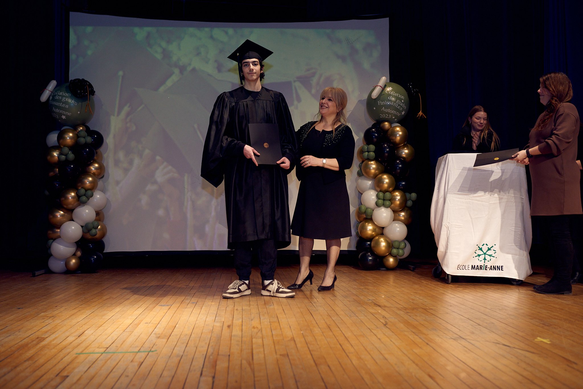 Une cérémonie de remise de diplôme à l'école Marie-Anne, avec une personne en tenue de diplômé, une autre en robe noire, deux autres personnes à l'arrière avec un pupitre. Décorations de ballons et écran en arrière-plan.