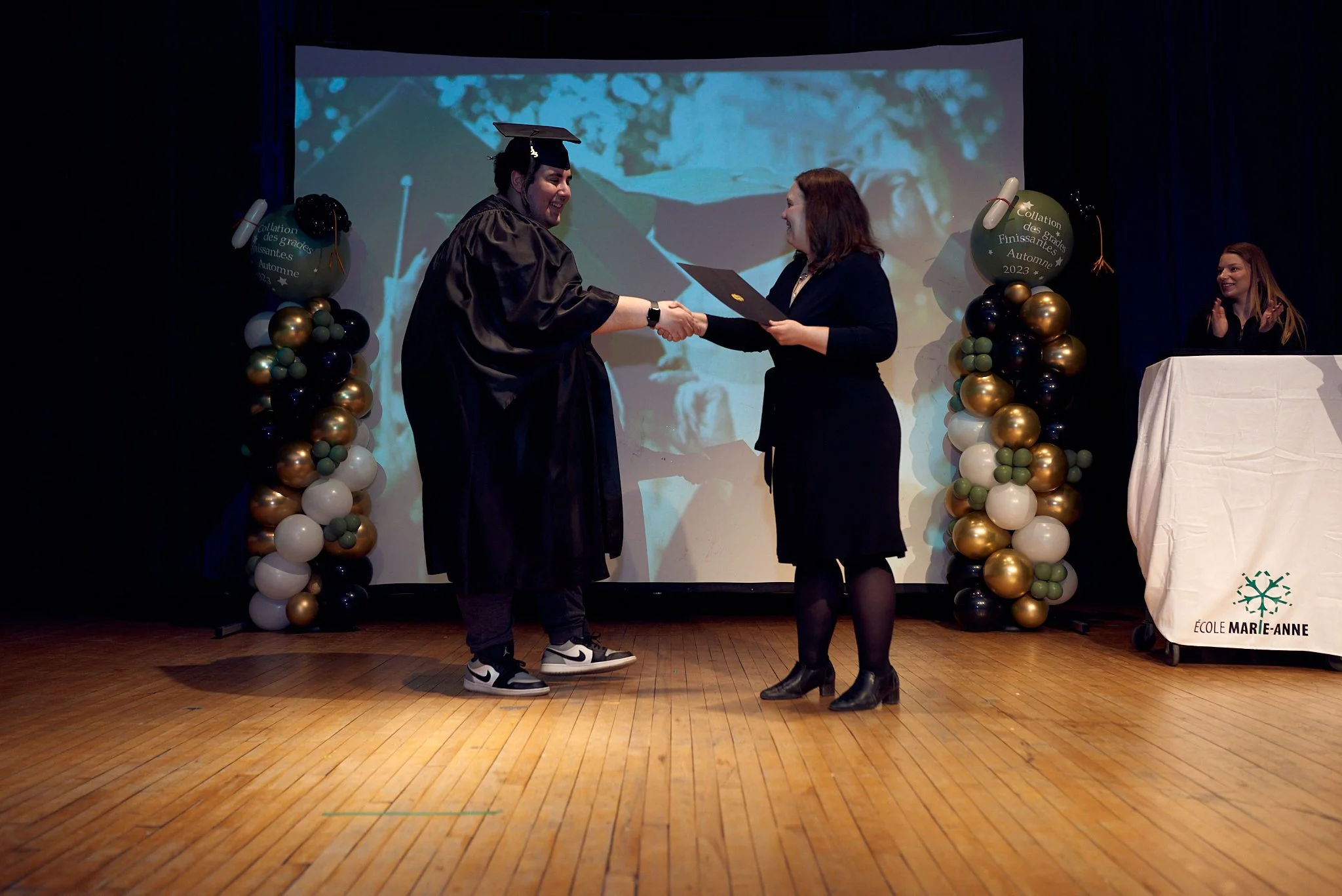 Une cérémonie de remise de diplôme sur scène, avec une personne en robe de graduation qui serre la main d'une autre personne, lors d'une cérémonie à l'école Marie-Anne.