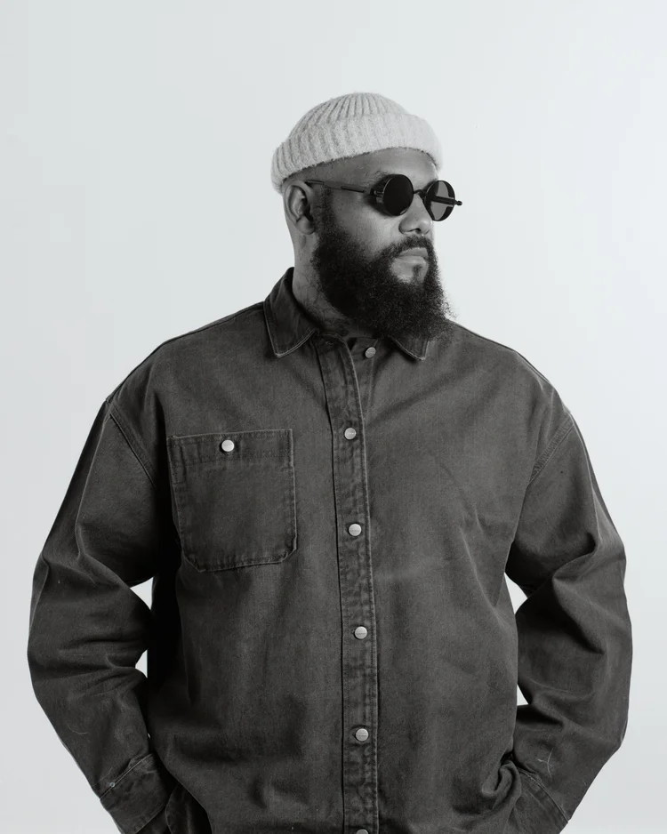 Homme portant un bonnet en laine, des lunettes rondes et une veste en denim sombre, posé de profil contre un fond blanc.