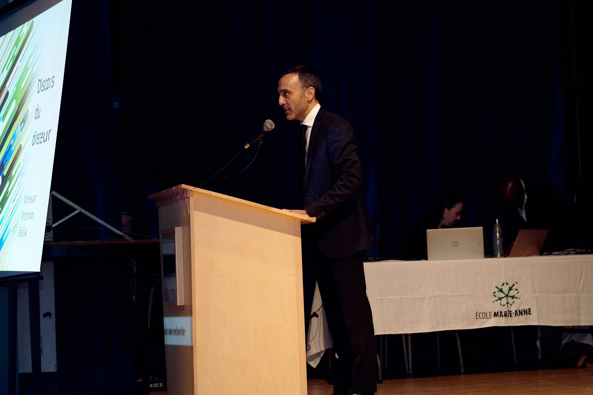 Homme en costume noir parlant derrière un podium lors d'une conférence, avec un écran à gauche et deux personnes en arrière-plan à une table.