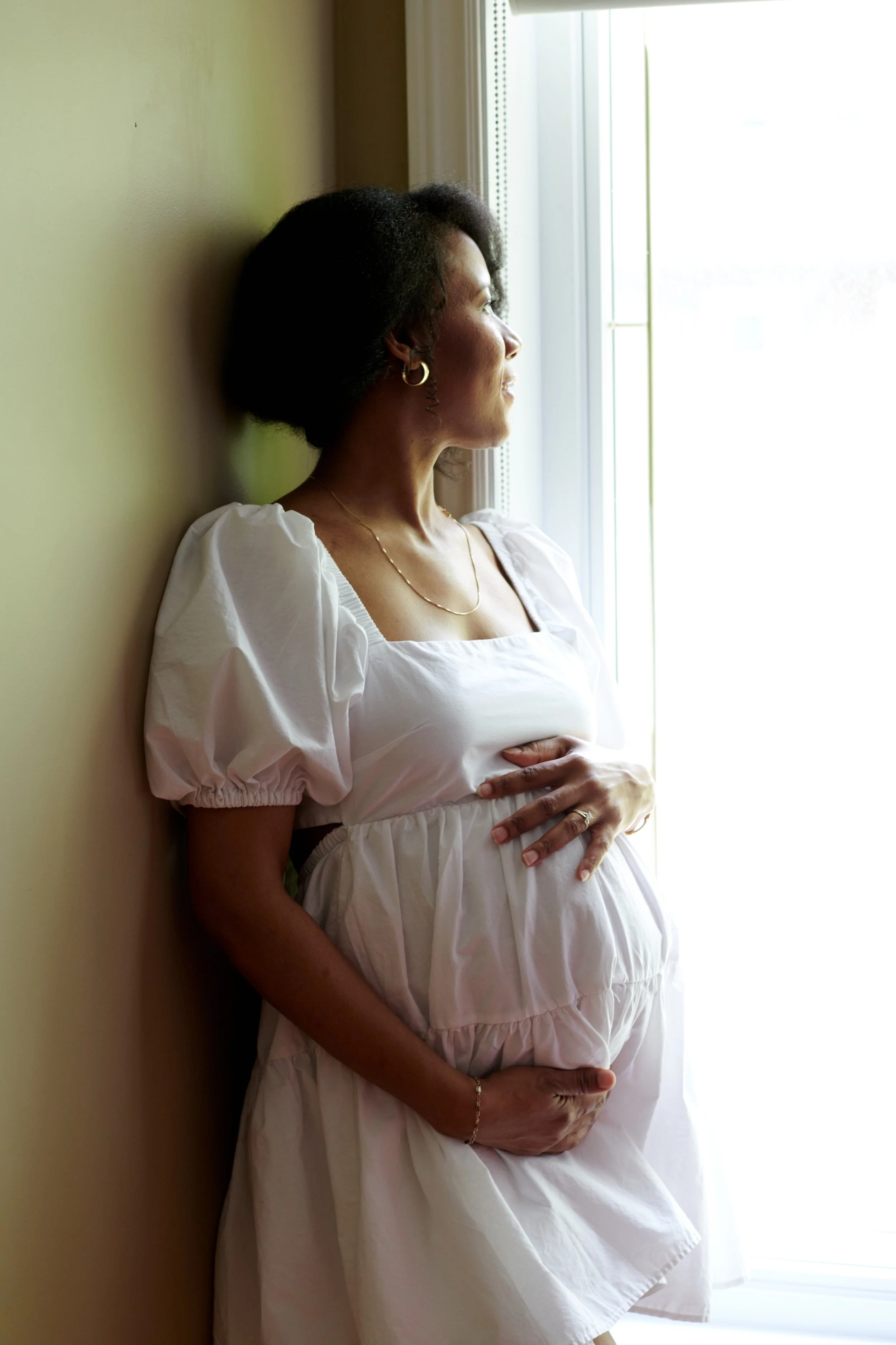 Femme enceinte regardant par une fenêtre, portant une robe blanche avec des manches bouffantes, tenant son ventre avec une main.