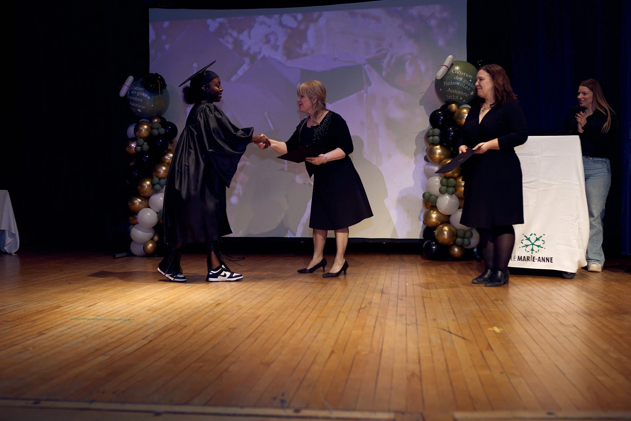 Une cérémonie de remise de diplôme lors d'une graduation, avec une étudiante en robe de graduation recevant un diplôme et serrant la main d'une femme en robe noire, sur une scène décorée de ballons.