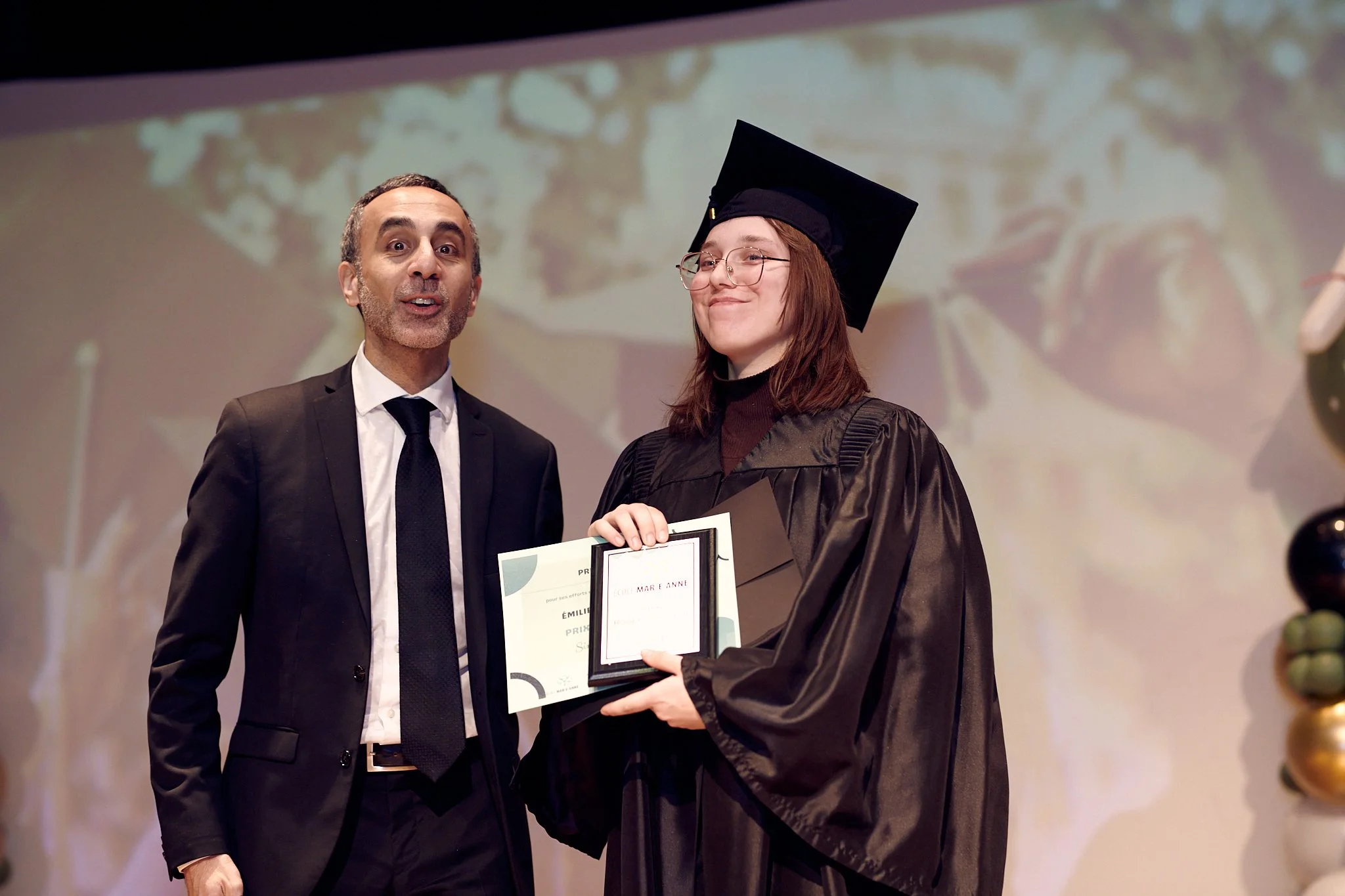 Une femme portant une toge et un chapeau de graduation recevant un diplôme lors d'une cérémonie, accompagnée d'un homme en costume.