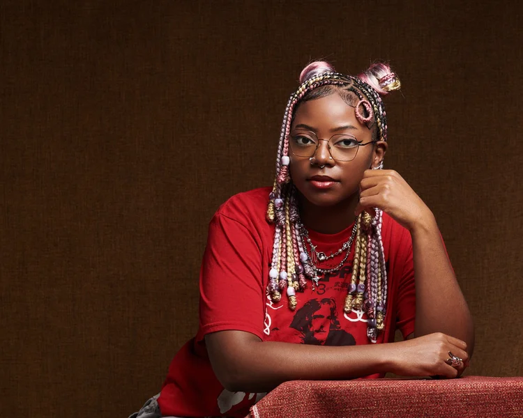 Jeune femme afro-américaine avec des cheveux colorés en tresses, portant des lunettes, une robe rouge et de nombreux colliers perlés, posant contre un fond brun