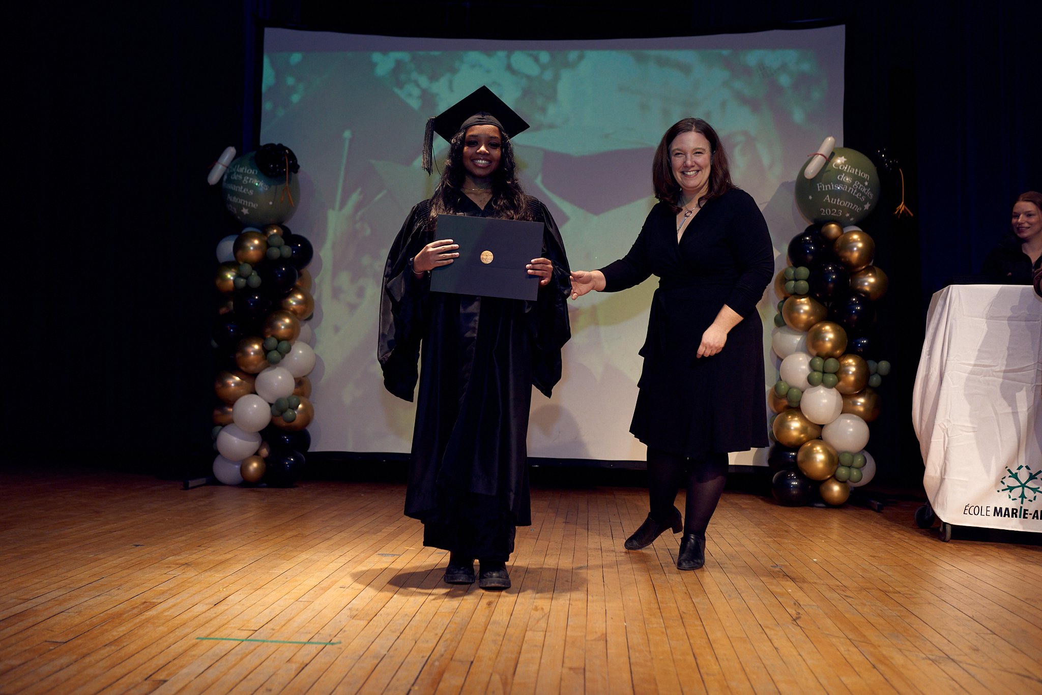 Une cérémonie de remise de diplôme d'études secondaires, une jeune fille en robe de graduation noire reçoit son diplôme d'une femme en robe noire, sur scène avec décor de ballons et un arrière-plan projette une image liée à l'automne.