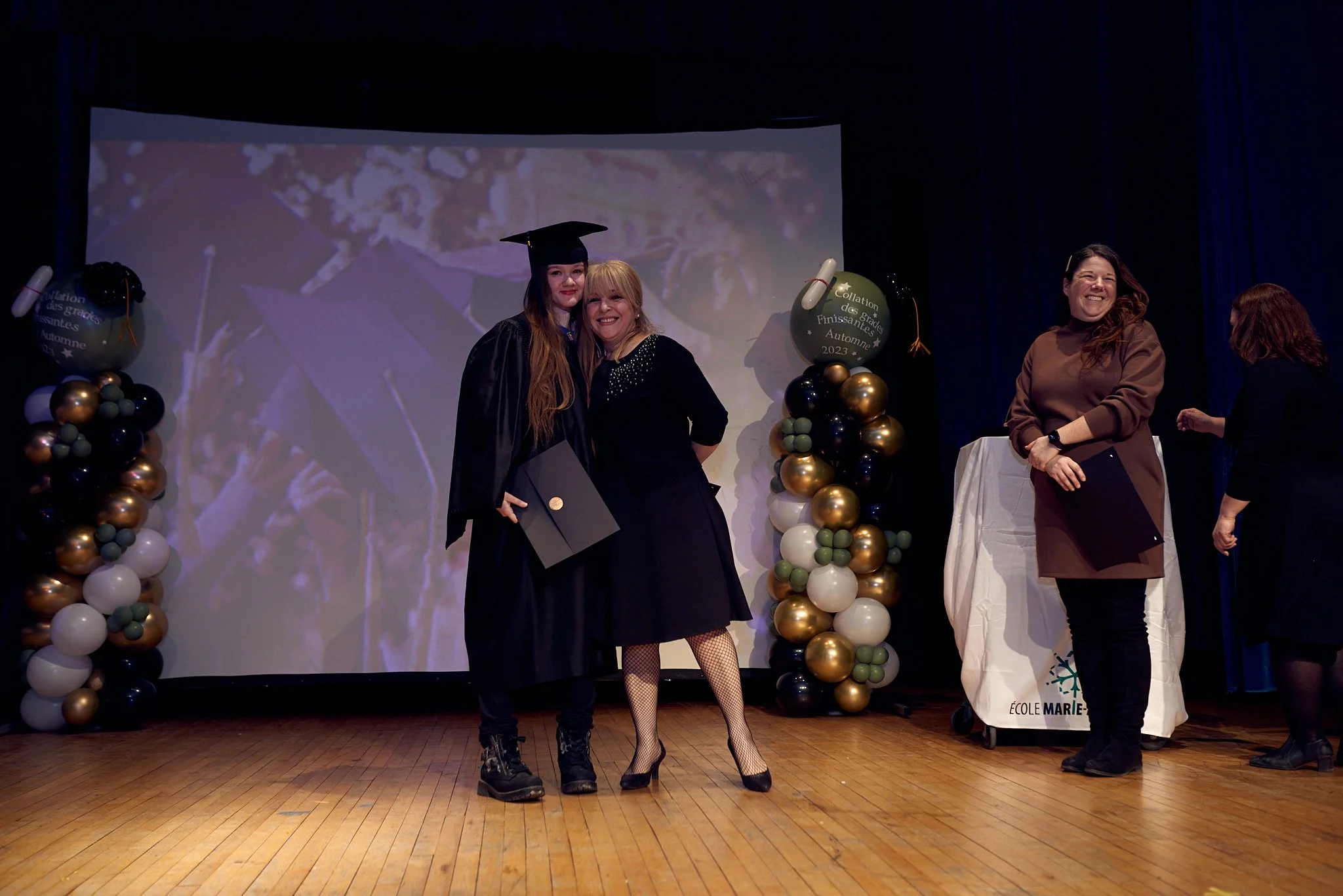 Une jeune femme en costume de graduation avec une robe noire, un chapeau de diplômé, tenant un diplôme, sourit aux côtés d'une femme en robe noire lors d'une cérémonie de remise de diplômes. Il y a des décorations avec des ballons noirs, blancs, et d