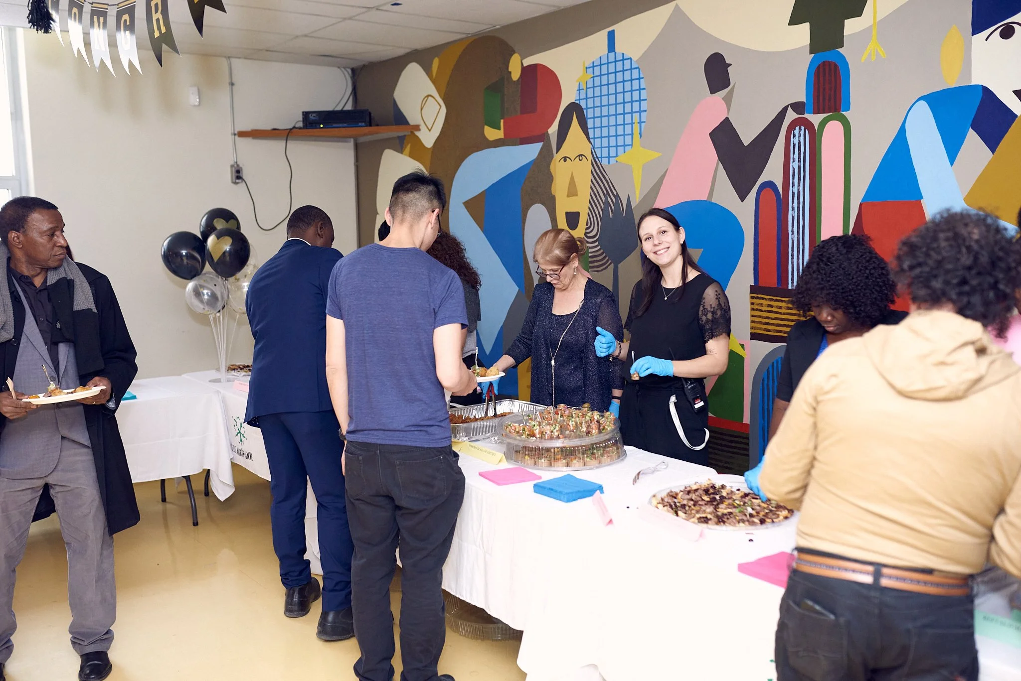 Groupe de personnes lors d'un événement social autour d'un buffet avec des bouchées et des boissons, dans une pièce décorée avec une fresque murale abstraite colorée.