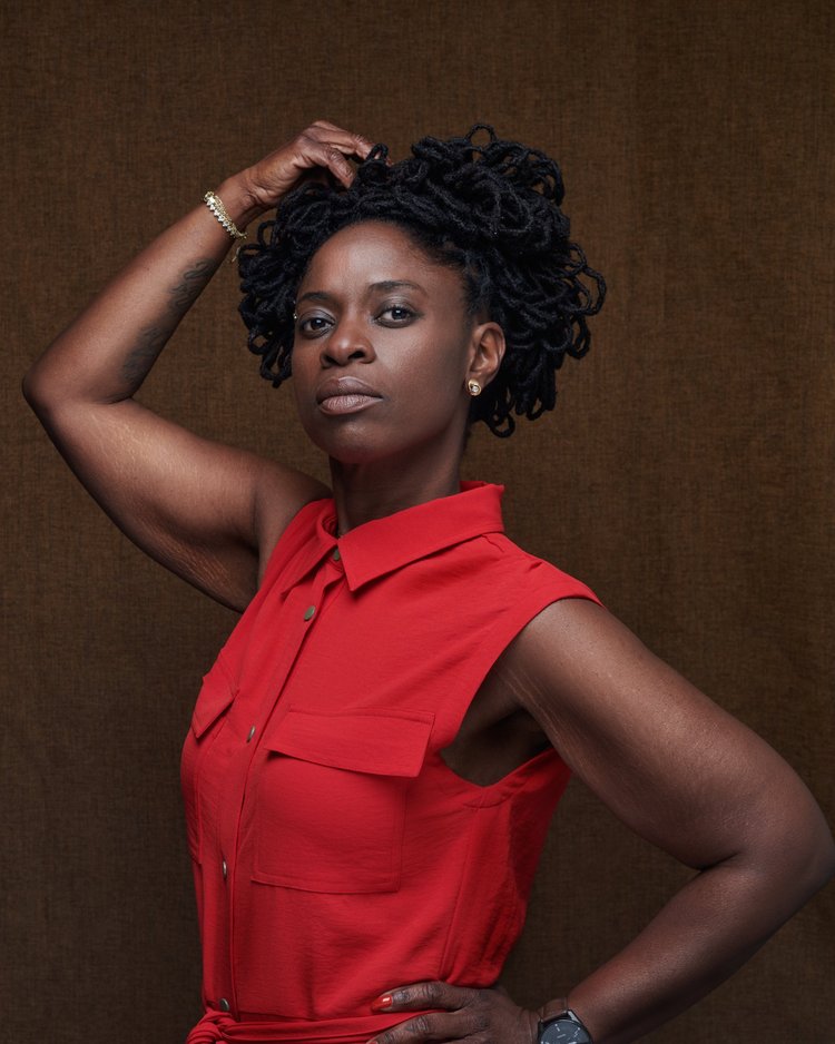 Portrait d'une femme noire portant une robe rouge, avec des cheveux en dreadlocks, en train de poser avec une main sur la tête et l'autre sur la hanche, contre un fond marron.