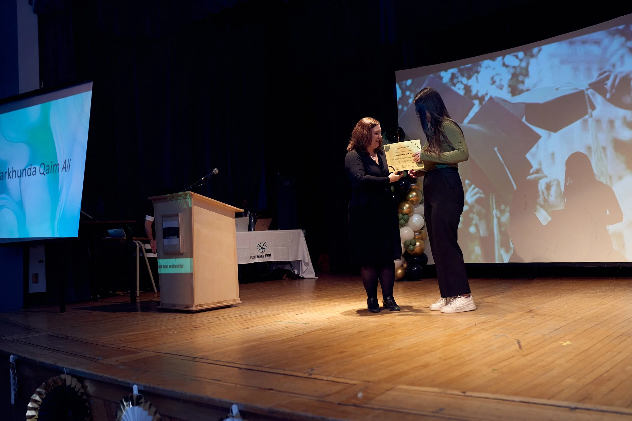 Une cérémonie de remise de diplôme ou de récompense sur scène, avec deux femmes échangeant un diplôme, l'une plus âgée et l'autre plus jeune, dans un auditorium avec un grand écran vidéo en arrière-plan.