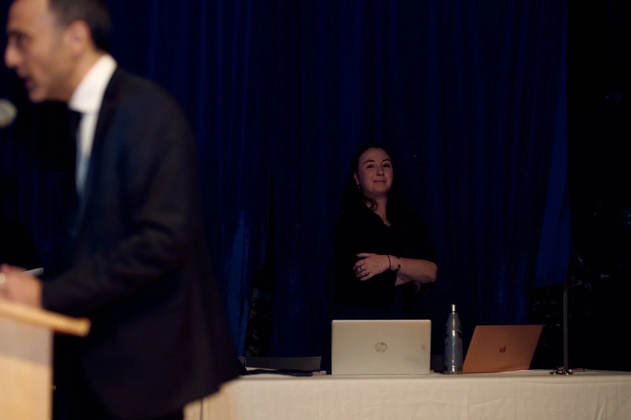 Une femme debout derrière une table avec deux ordinateurs portables, en fond une scène avec des rideaux bleus et un homme en costume blanc et noir flou à gauche.