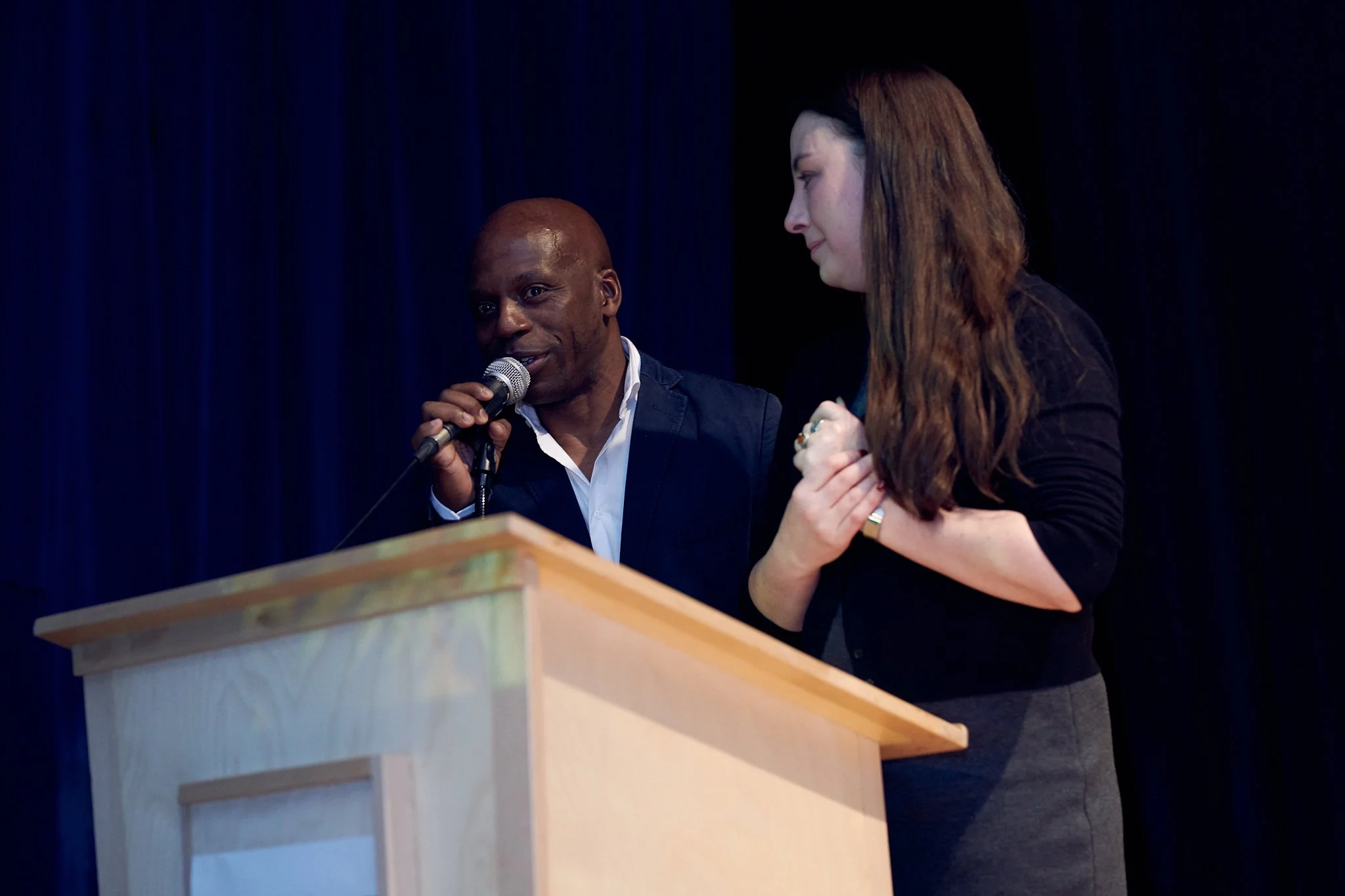 Deux personnes debout derrière un podium, l'une parlant dans un microphone, l'autre en train de la regarder avec attention, sur scène avec un fond sombre.